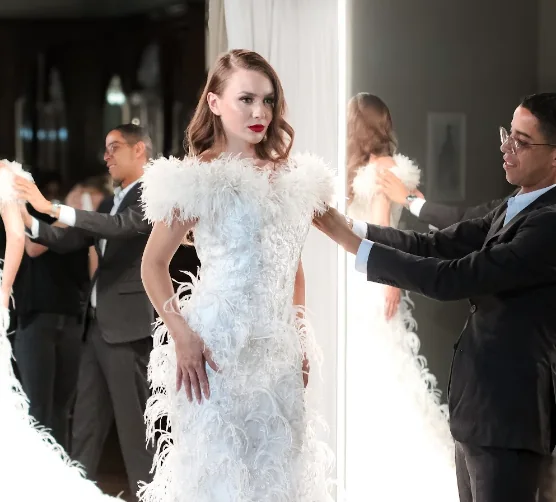 Leandro Fonseca ajustando um vestido de plumas na modelo, diante do espelho.