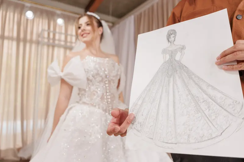 O croqui a lápis na mão do estilista; ao fundo, desfocada, a noiva veste o próprio vestido pronto.